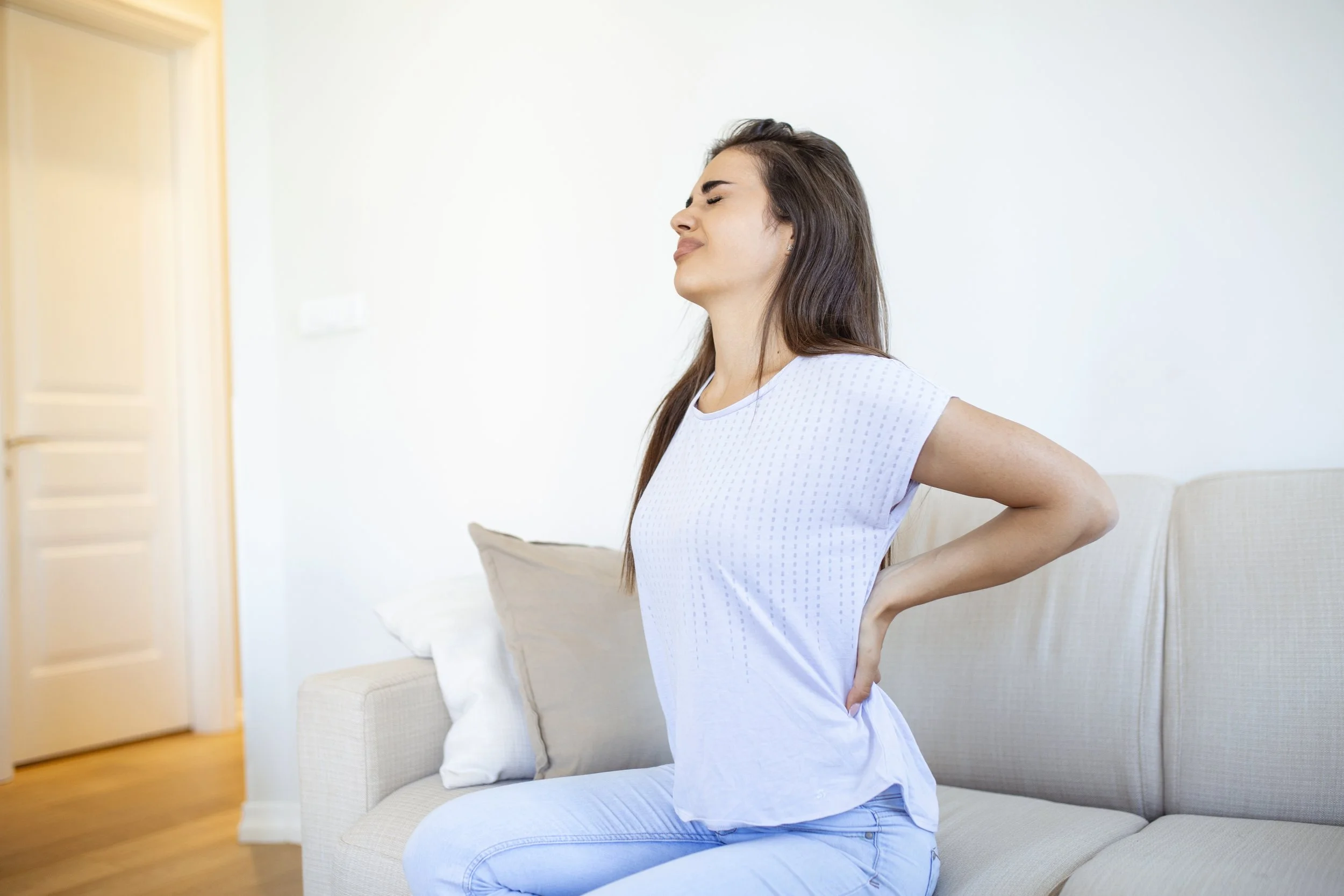 A woman sitting on a sofa clutching her mid-back in pain, seeking specialized physical therapy for scoliosis and spine deformation at Signature PT.