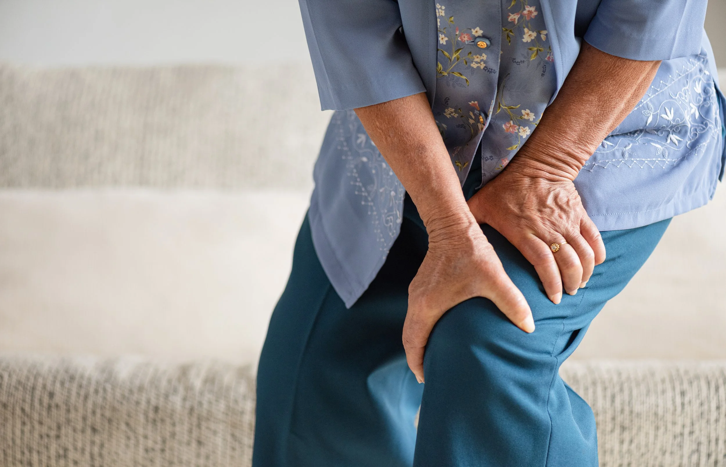 An elderly woman in blue clothing clutching her knee while trying to stand up, illustrating symptoms of osteoarthritis and functional mobility loss.
