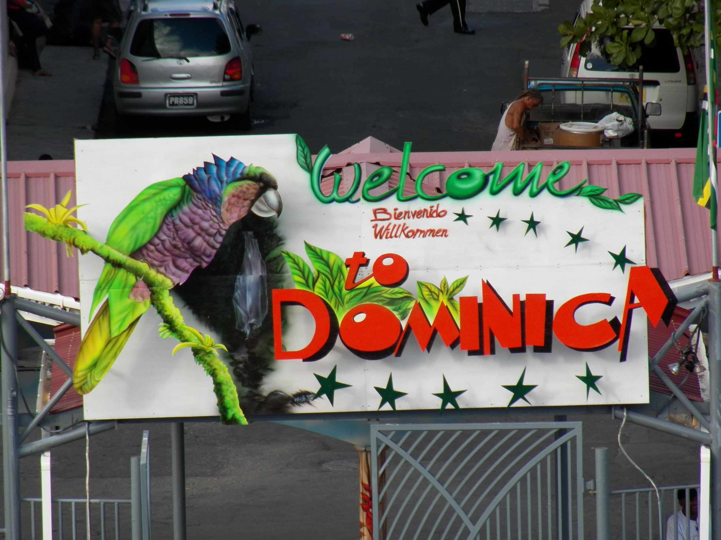 A colorful sign with a parrot illustration and the words 'Welcome' in green, 'Bienvenido', 'Willkommen' in red, and other decorative elements. In the background, there is a parking lot with cars and a person standing near a truck.