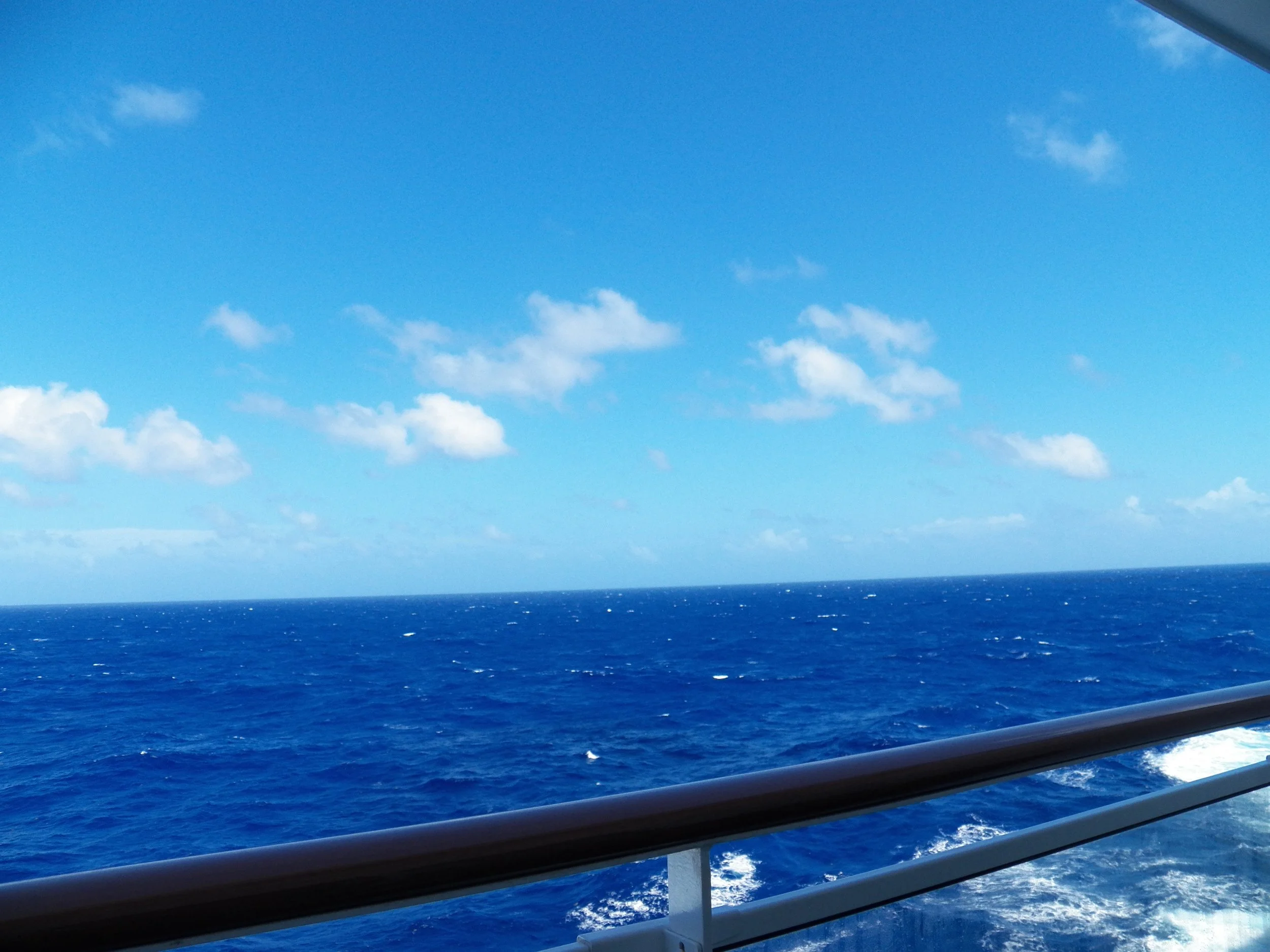 View of the ocean with a deep blue sea and a bright blue sky with some scattered clouds, taken from a boat or a ship.