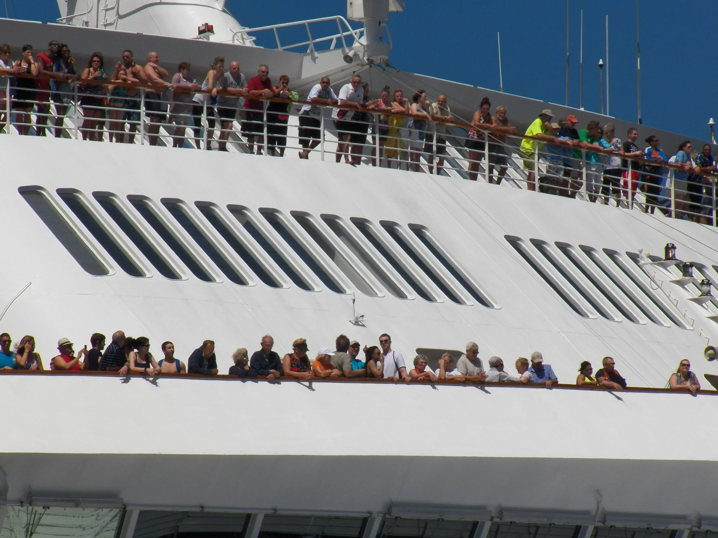 Crowd of people standing on upper deck of cruise ship, enjoying sunny weather and sightseeing.
