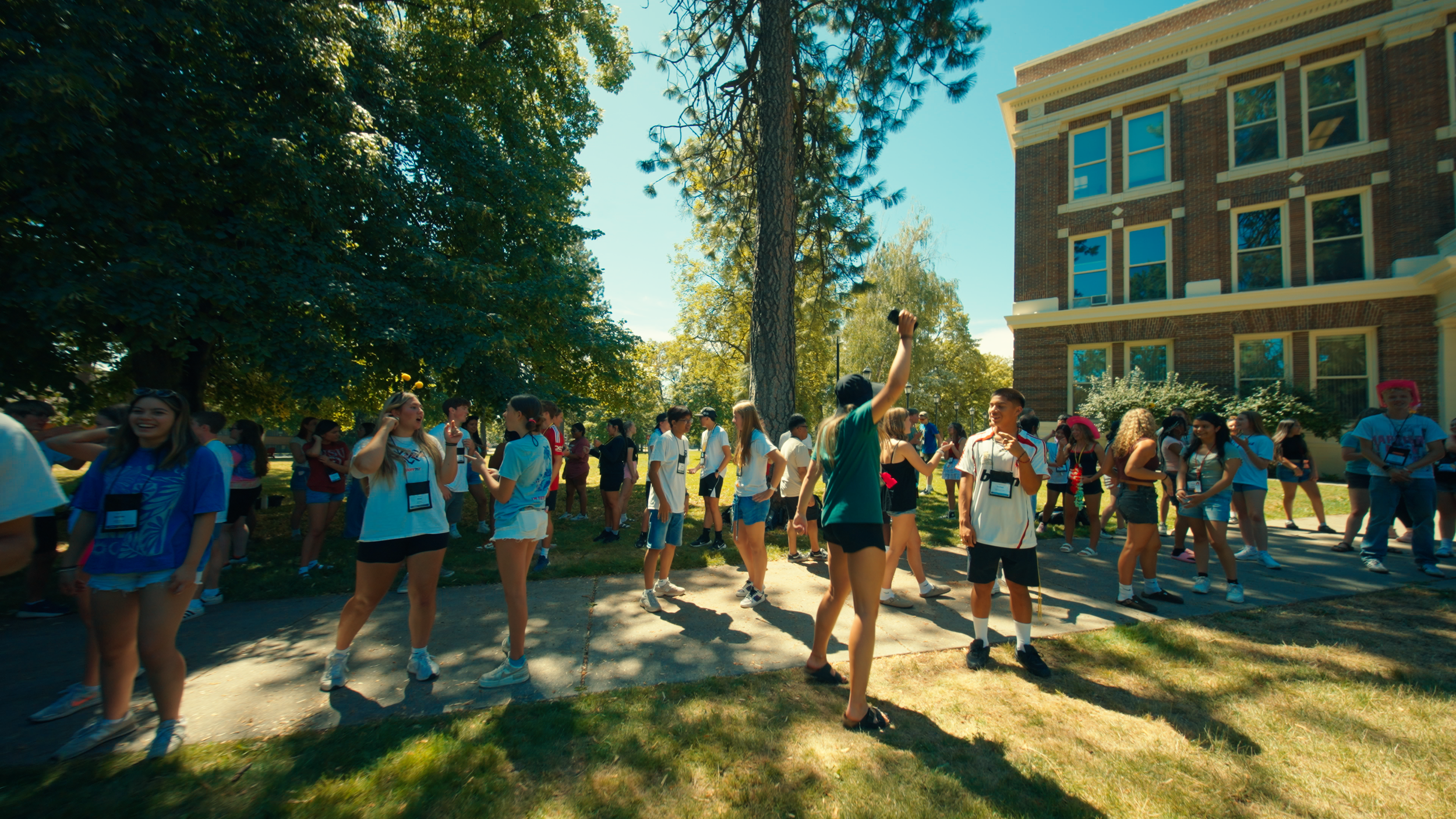 A group of young people outdoors on a sunny day, socializing and smiling, with some wearing lanyards, under trees and near a brick building.