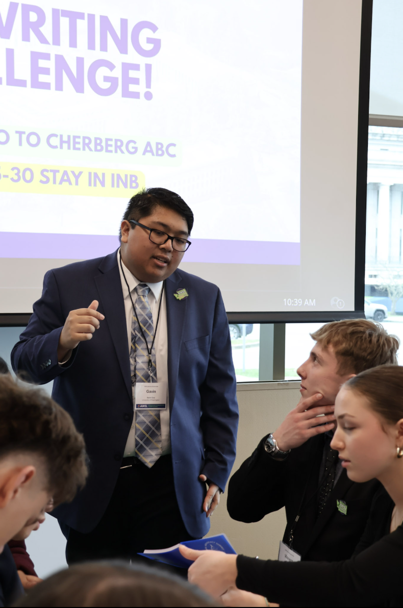 A man in a blue suit and glasses standing and speaking to a group of people seated around a table. A presentation slide is visible in the background with the words 'Writing Challenge!' and additional text, along with a digital clock showing 10:39 AM.