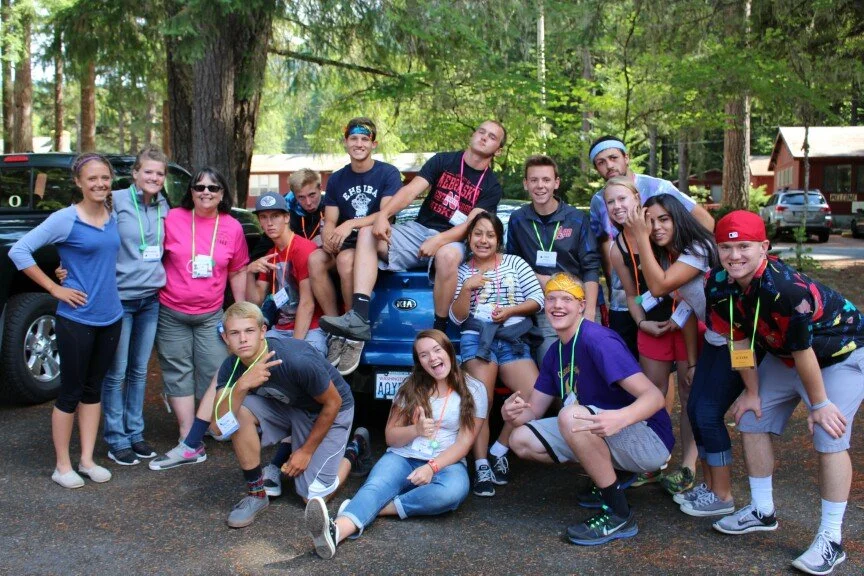 A group of teenagers and young adults posing outdoors in a wooded area, some sitting on and around a blue car, with everyone smiling and making playful gestures.