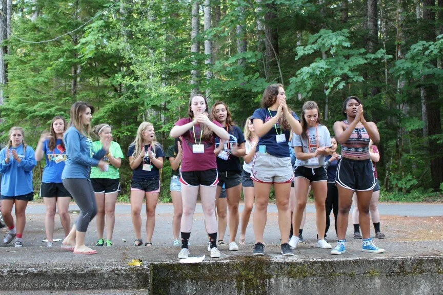 A group of young girls standing outdoors on a paved area, surrounded by green trees, with some of them holding hands in a prayer or meditative pose.