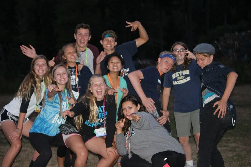 Group of kids and teenagers outside at night posing playfully for a photo, smiling and making silly gestures.