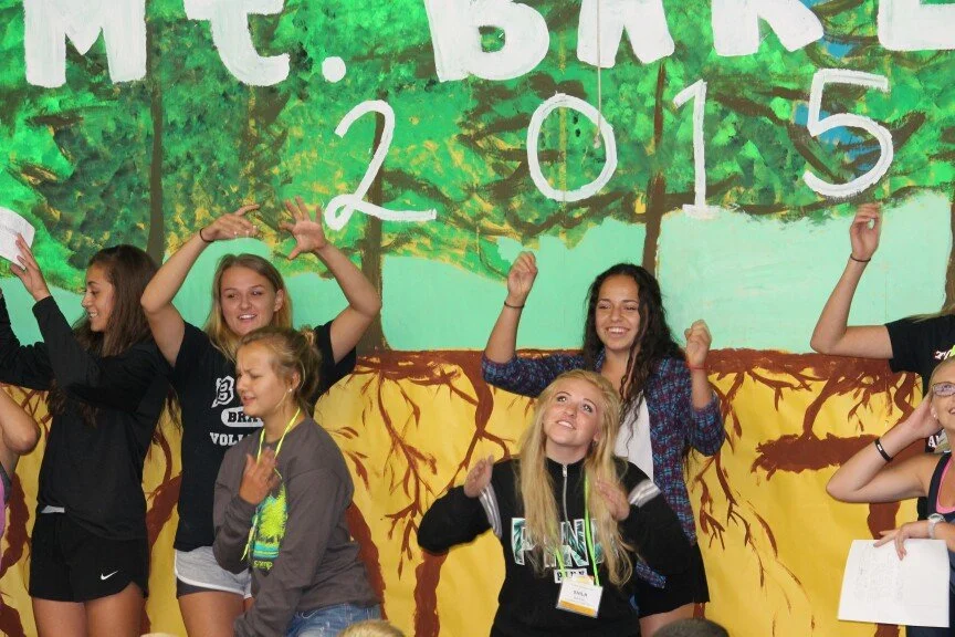 Group of smiling girls celebrating at a school event in front of a colorful backdrop with trees and the year 2015.