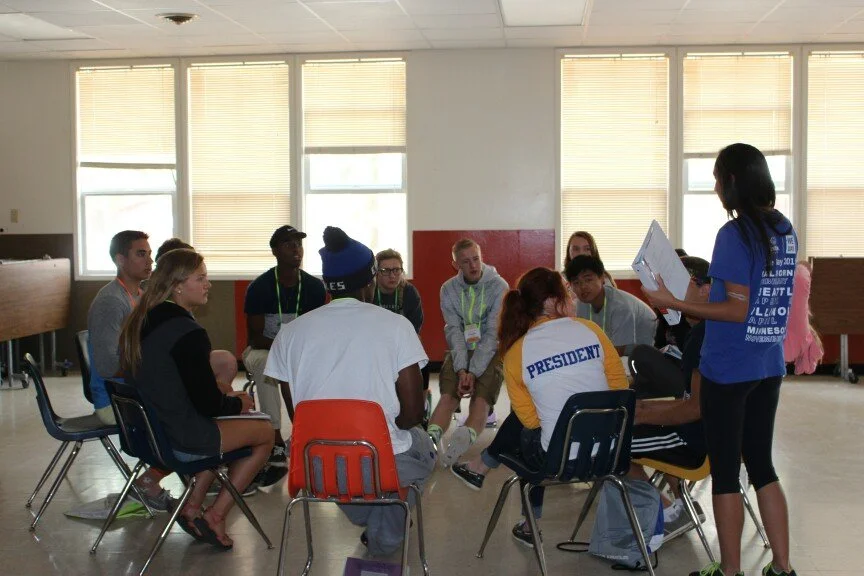 A group of young people seated in a circle on chairs in a bright room, listening to a woman in a blue shirt who is standing and holding papers, leading a discussion.