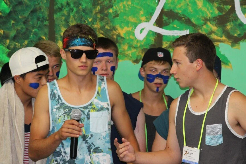 Group of boys, some wearing face paint and casual summer clothing, standing outdoors with a painted backdrop of trees and a white structure.