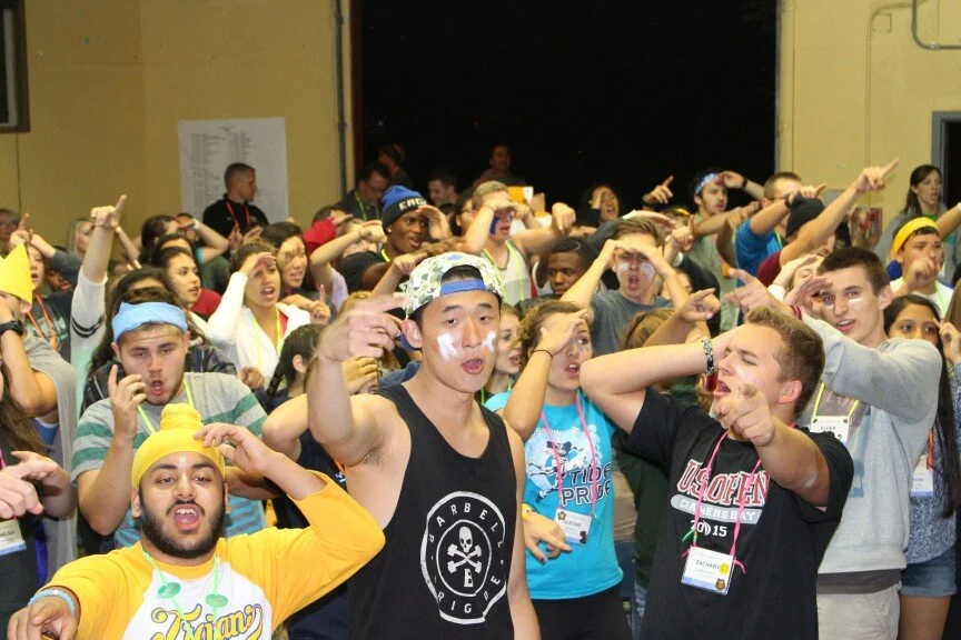 A large group of young people at an indoor event, raising their hands and pointing, some with face paint, wearing casual clothing and badges.