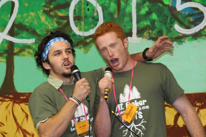 Two young men singing into microphones on a stage, wearing matching t-shirts with trees and roots graphic, with a colorful painted backdrop of trees in the background.