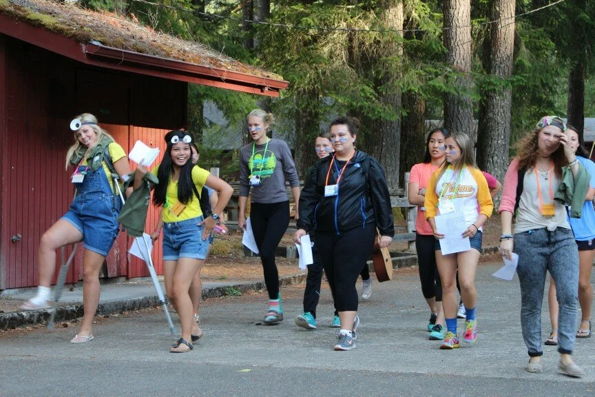 Group of young girls walking outdoors in a forested area, some wearing casual clothes and face paint, carrying papers and backpacks, enjoying a camp or outdoor activity.