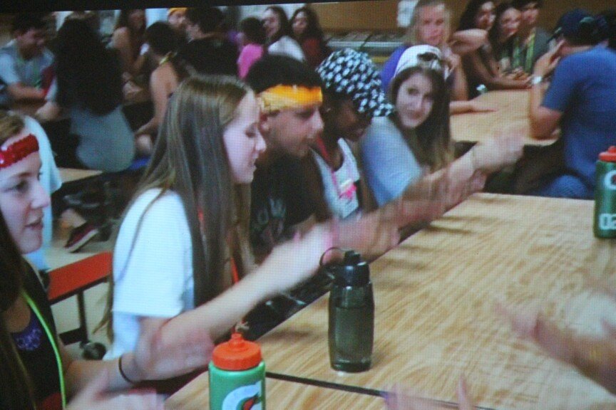 Group of young people sitting at a long wooden table in a crowded room, some wearing headbands and exploring their phones.