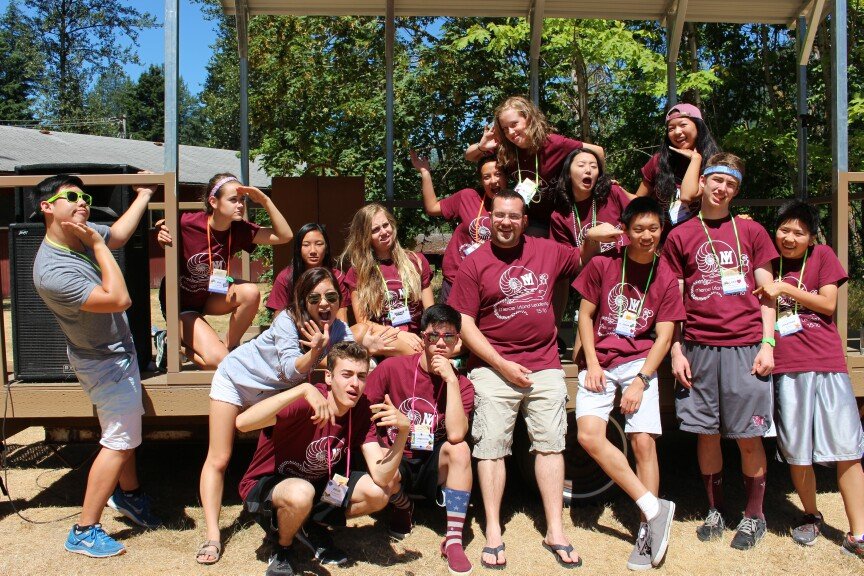 Group of teenagers and a man posing on an outdoor stage with trees in the background. Everyone is smiling and wearing casual clothes, some with event badges around their necks.