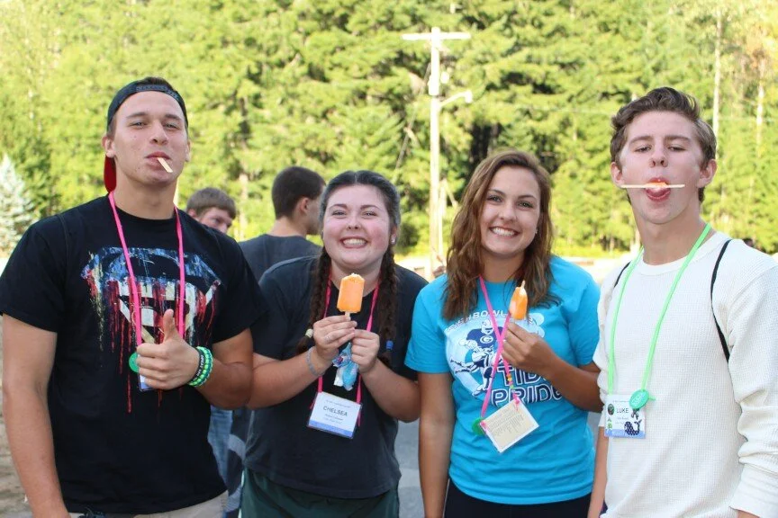 Four teenagers smiling and enjoying ice pops outdoors during daytime, standing together with a background of trees and some other people.