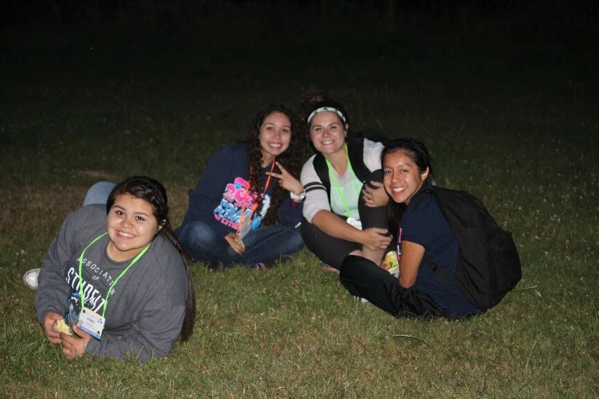 Four young women sitting on grass outdoors at night, smiling at the camera, with backpacks and name tags.