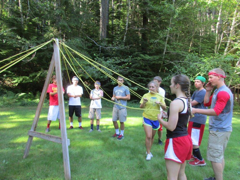 Group of children and teenagers participating in an outdoor team-building activity with a wooden pyramid and multiple yellow ropes in a grassy field surrounded by trees.