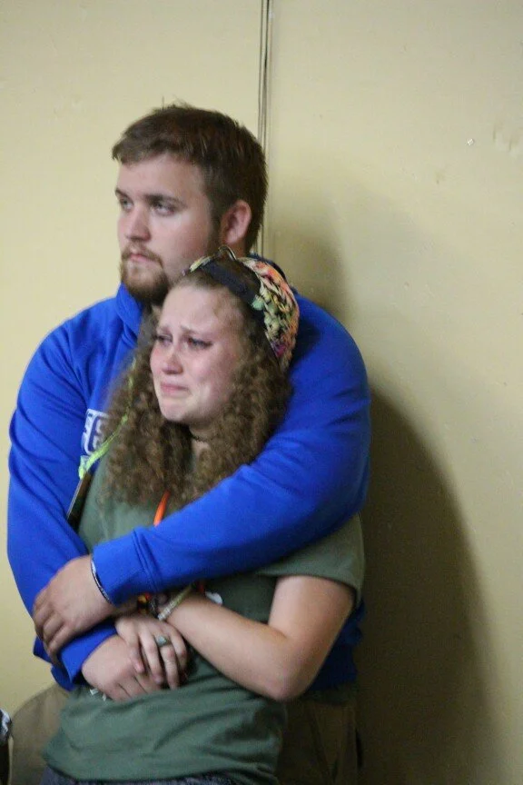 A young man with a beard wearing a blue hoodie hugs a young woman with curly hair, a floral headband, and a gray t-shirt, who appears distressed and is holding her arm.