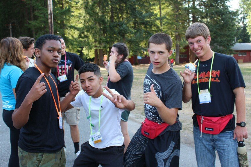 Four teenage boys standing outdoors in a park, making gestures towards the camera, some with lanyards and name tags, others holding food items, with trees and a building in the background.