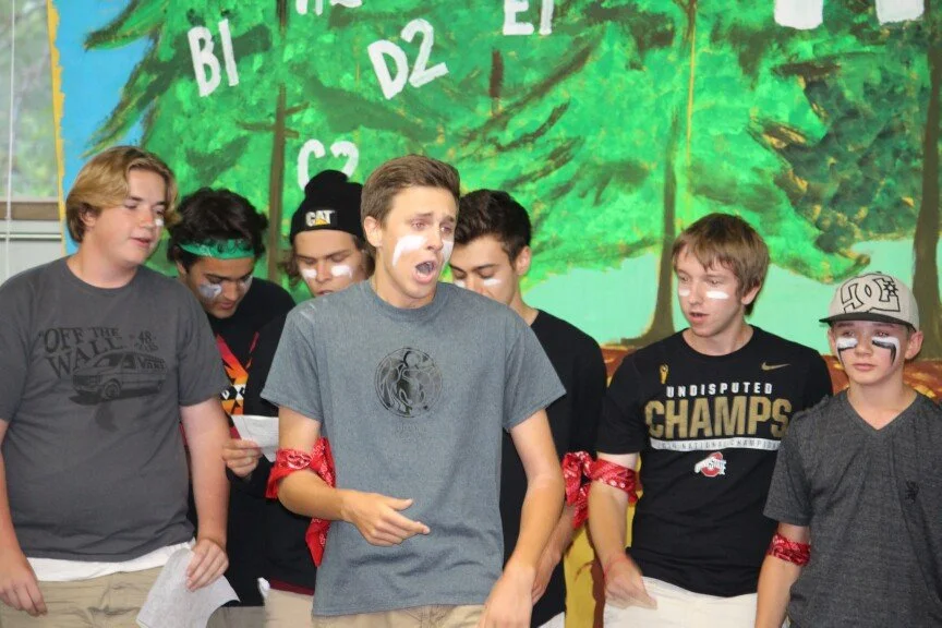 A group of young boys standing in front of a painted mural of trees and letters. They are wearing casual clothes, with some having face paint and red bandanas on their arms, participating in an indoor activity or performance.