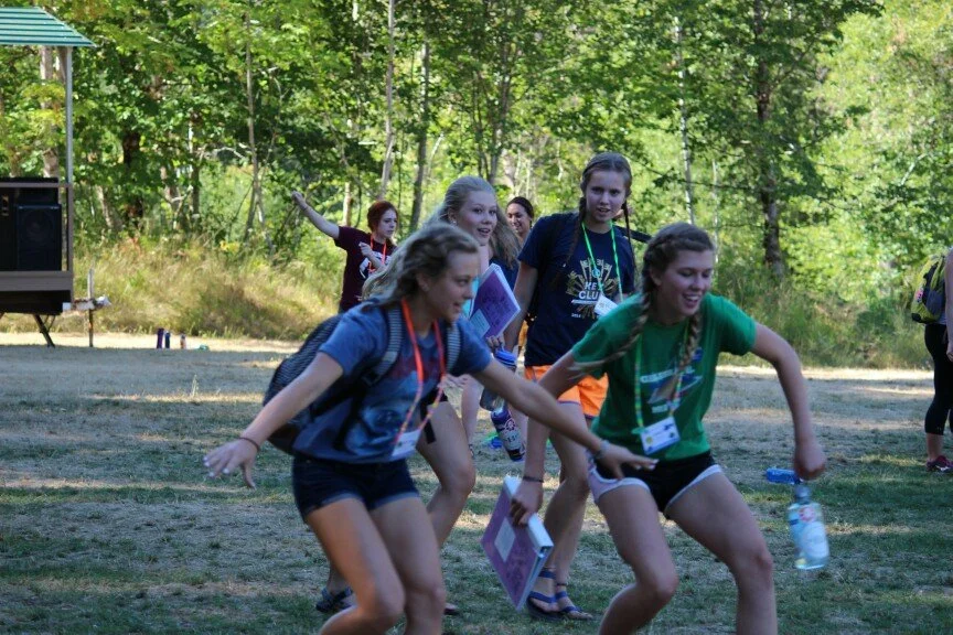 Group of girls at an outdoor summer camp or retreat, holding water bottles and notebooks, playing on grassy field with trees in the background.
