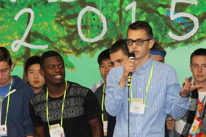 Group of diverse students at a school event, with a background of painted trees and the year 2015, some wearing name tags, and one student speaking into a microphone.