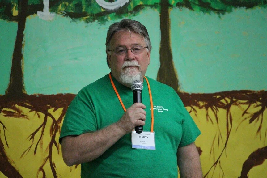A man with gray hair, glasses, and a beard holding a microphone, wearing a green shirt with a name tag, standing in front of a colorful mural of trees and roots.