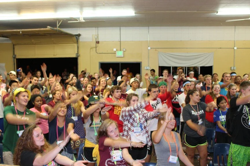 Large group of young people dancing and raising their hands inside a spacious hall.