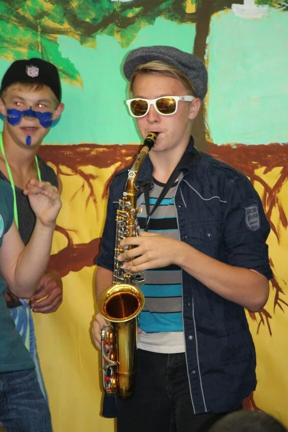 A boy wearing sunglasses and a hat is playing a saxophone, standing in front of a painted background with trees. Another boy with face paint is partially visible beside him.