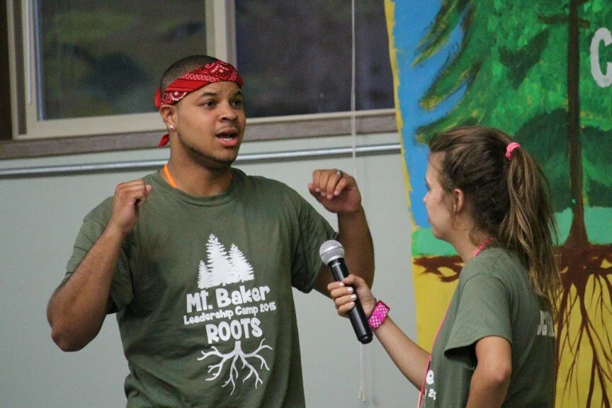 A young man with a red bandana speaking passionately during an interview or discussion, held by a young girl holding a microphone, both wearing green T-shirts at a leadership camp, with a colorful mural in the background.