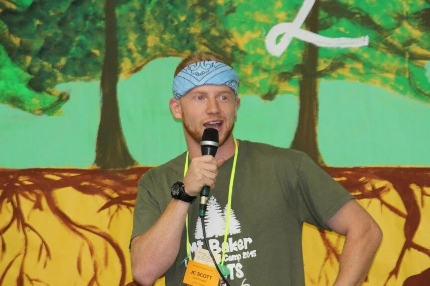 A man wearing a blue bandana and a green T-shirt with a logo, speaking into a microphone at an outdoor event with a colorful forest-themed backdrop.