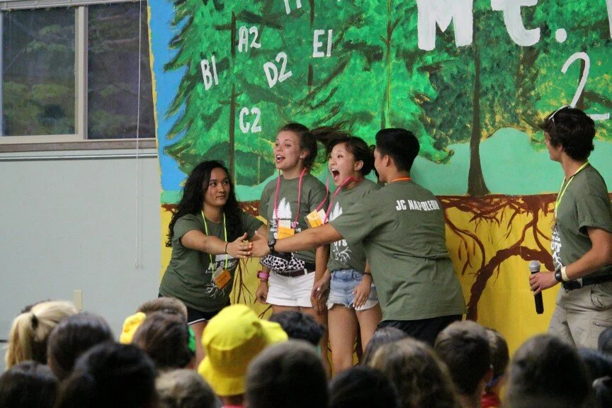 Group of young people acting in a skit or play on stage in front of an audience, with a colorful painted tree backdrop.