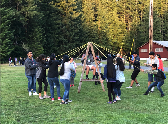 Group of people outdoors playing a tetherball game with a wooden A-frame structure, surrounded by trees.