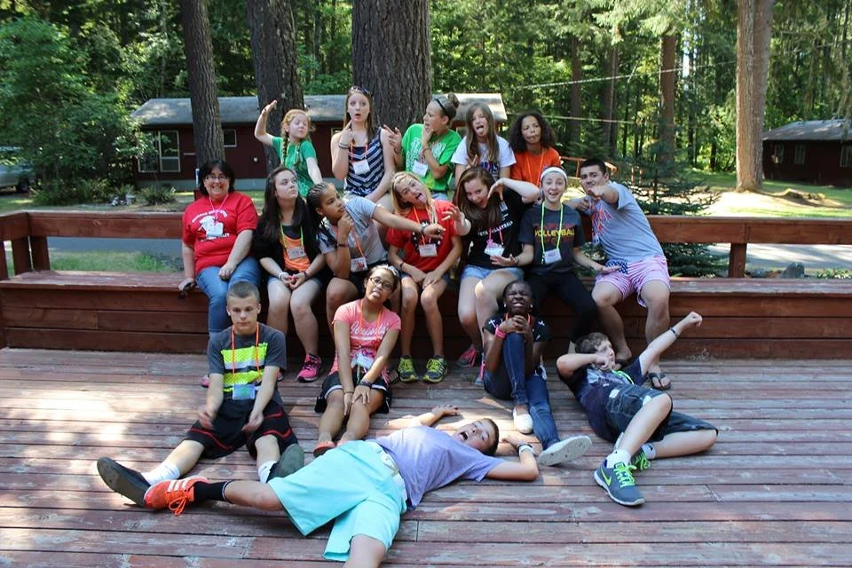 A group of children posing and making silly faces on a wooden deck outdoors surrounded by trees.