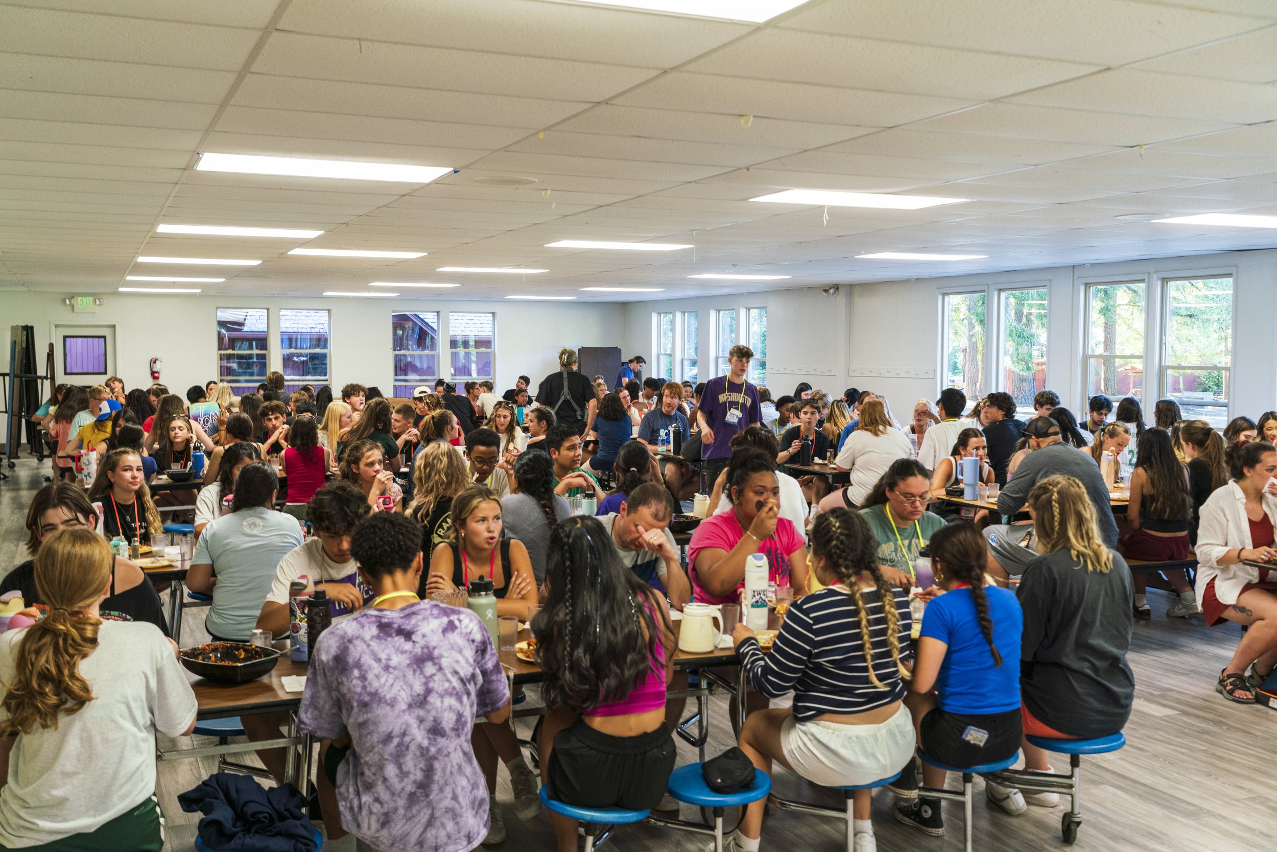 A large group of young people sitting at long tables in a bright, spacious room with large windows, eating and socializing.