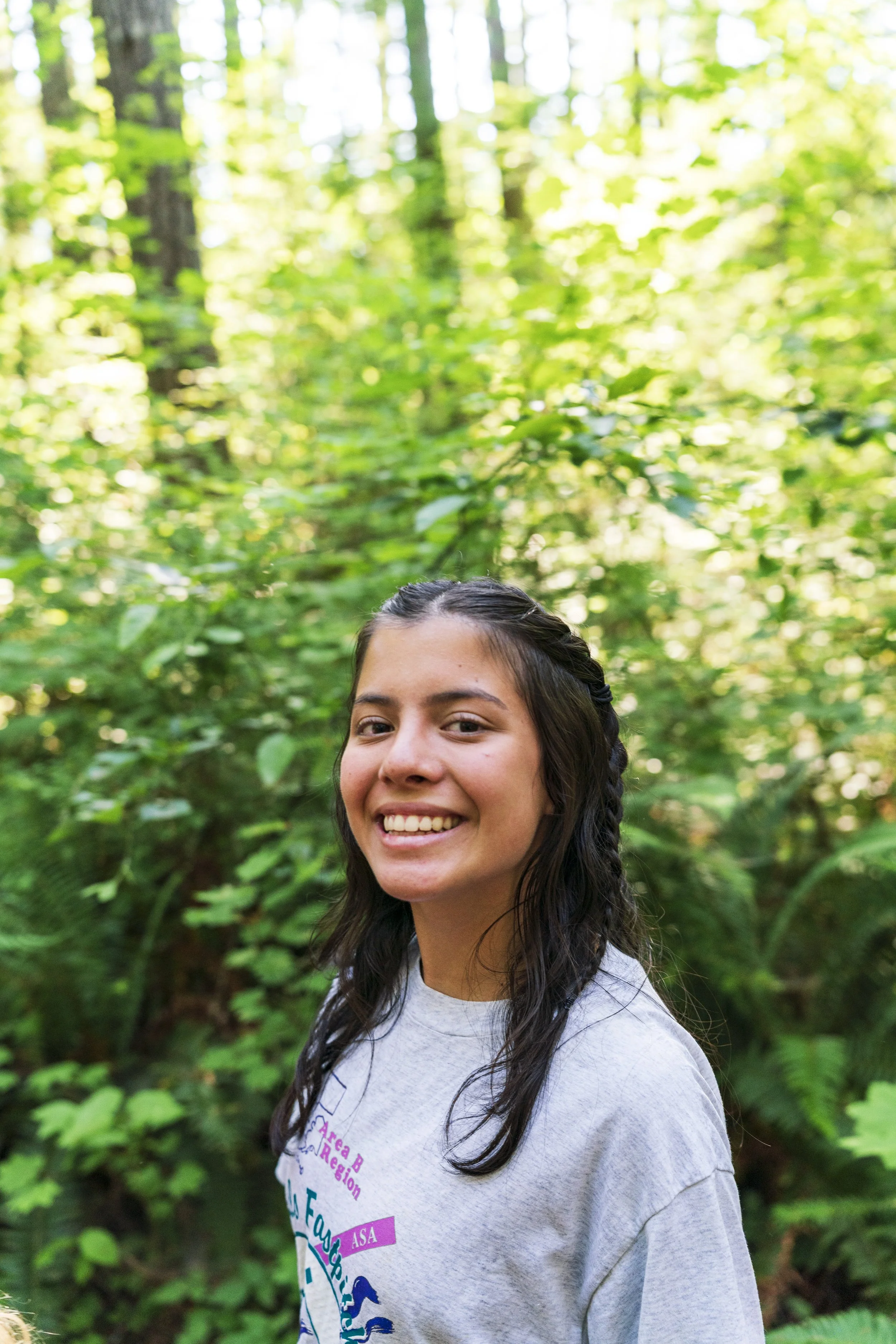 A smiling woman with long dark hair in a braid, wearing a light gray T-shirt, standing in a lush green forest.