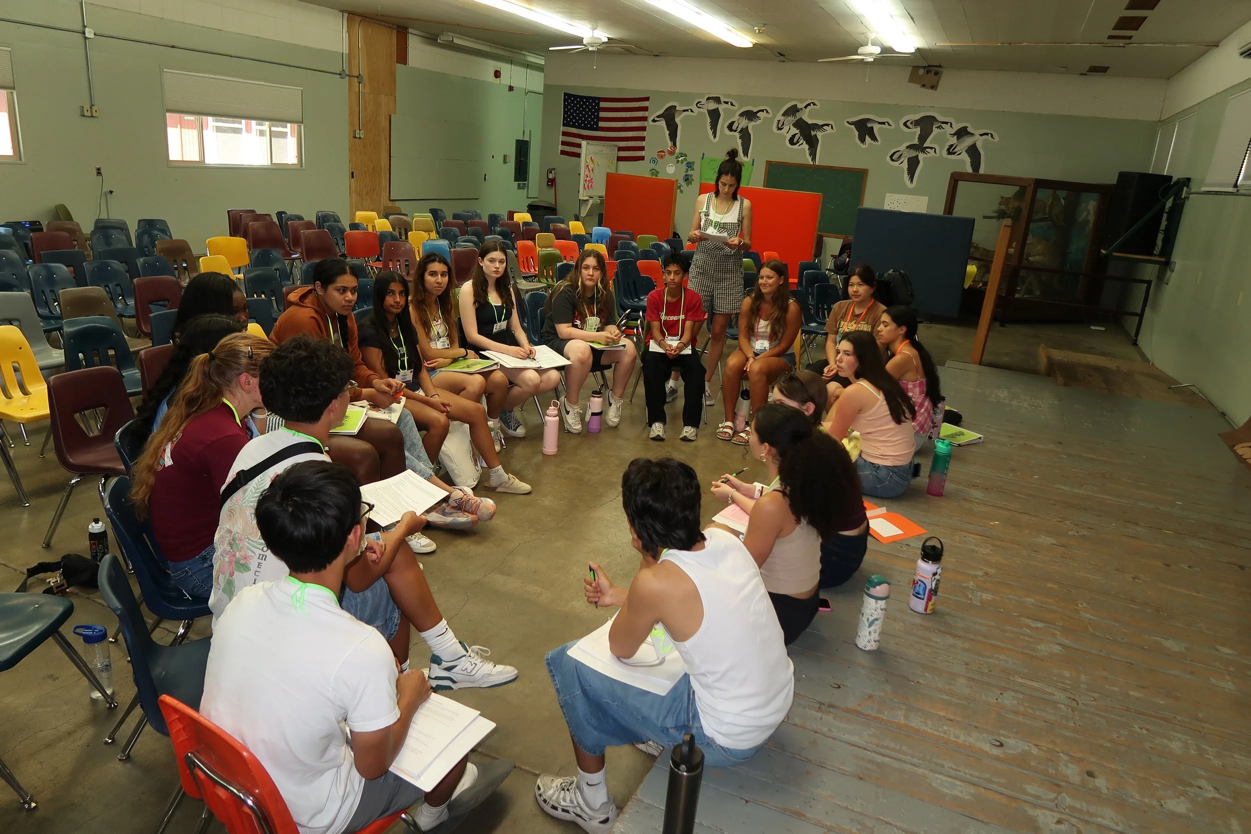 A group of young students, mostly female, seated in a circle in a classroom with colorful chairs, listening to a woman standing and speaking. The classroom has American flag and bird decorations on the wall.