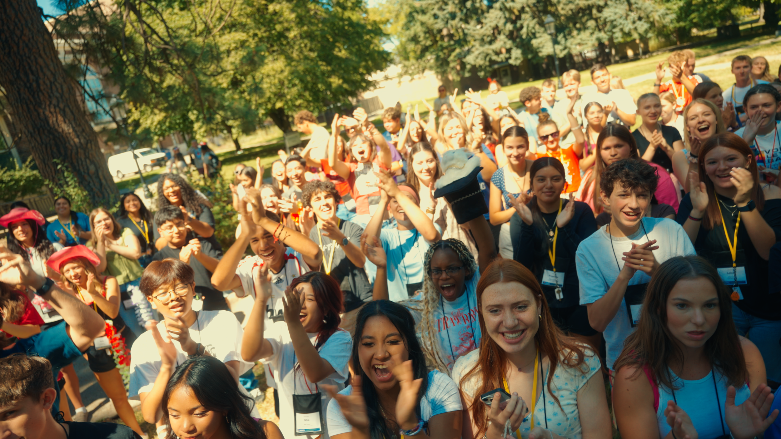 Large group of diverse children and teenagers outdoors in a park, smiling and cheering, some wearing name tags and colorful clothes, with trees and sunlight in the background.