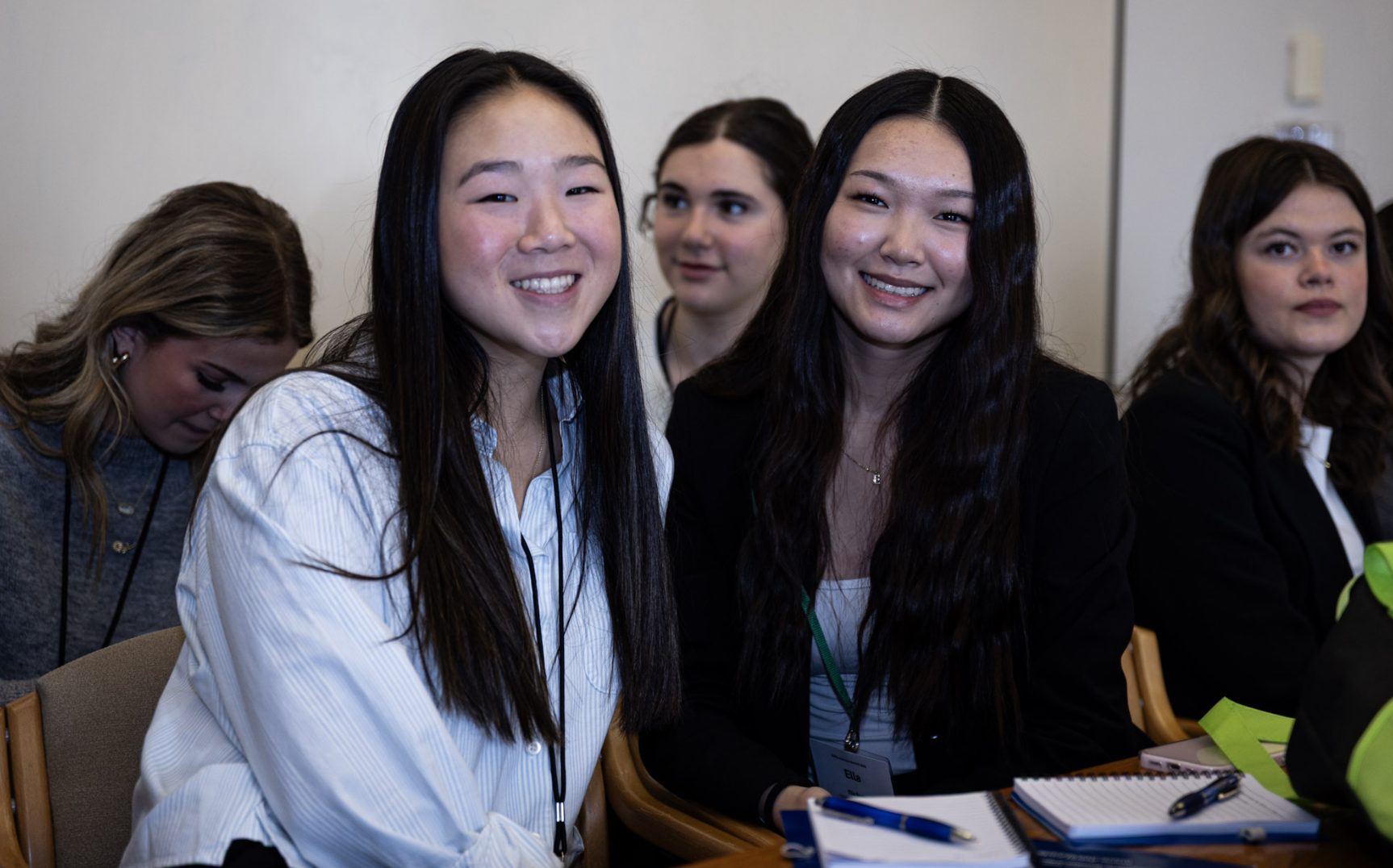 Group of five young women sitting at a conference table, smiling and looking towards the camera, with notebooks and pens in front of them.