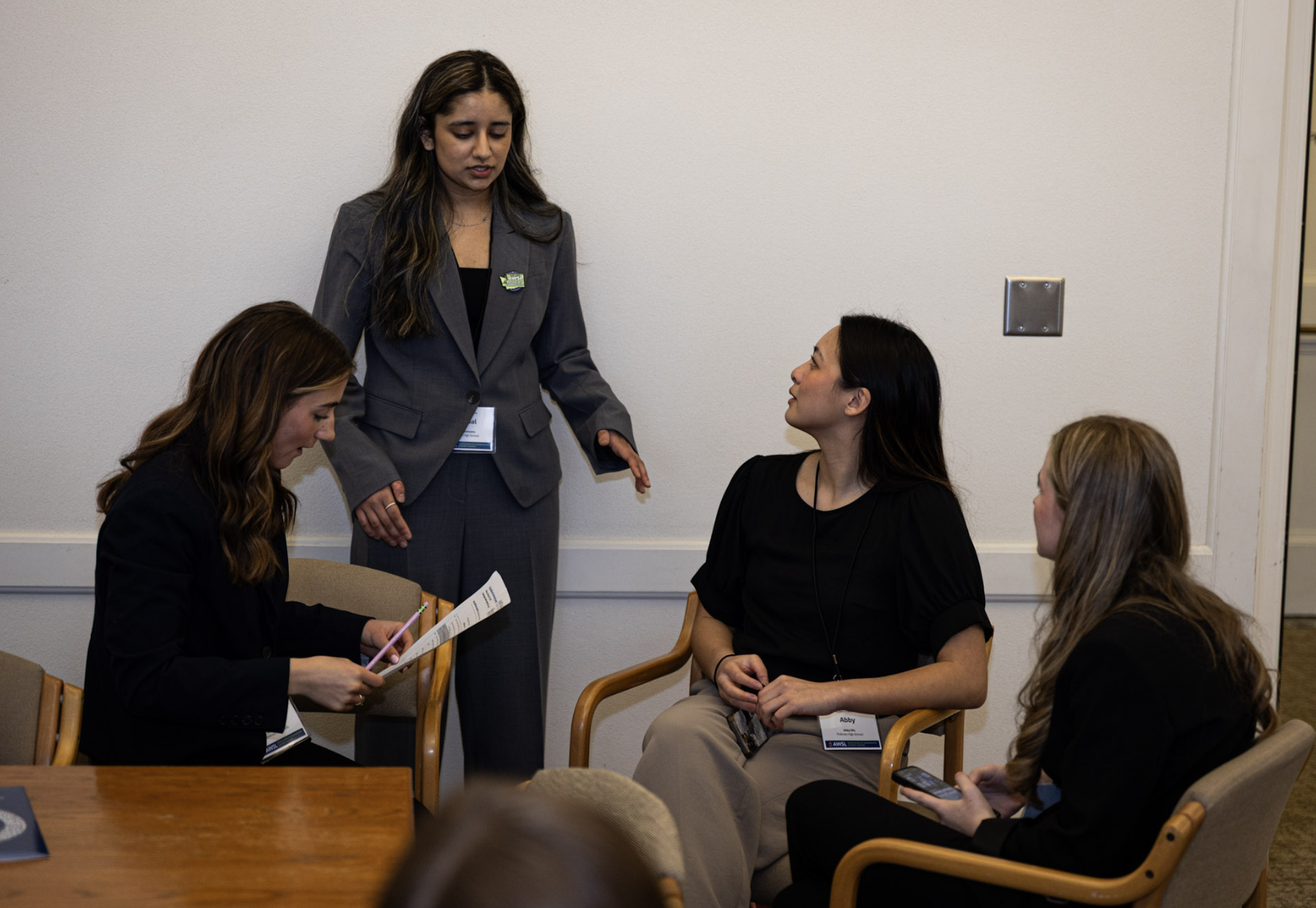 Five women are engaged in conversation during a professional meeting or conference in a room with beige chairs and light-colored walls. Four women are seated, while one woman is standing, talking to the group.