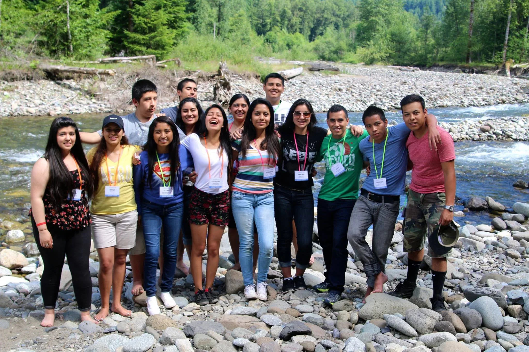 Group of young people standing barefoot on rocky riverbank in a forested area, smiling and posing together.