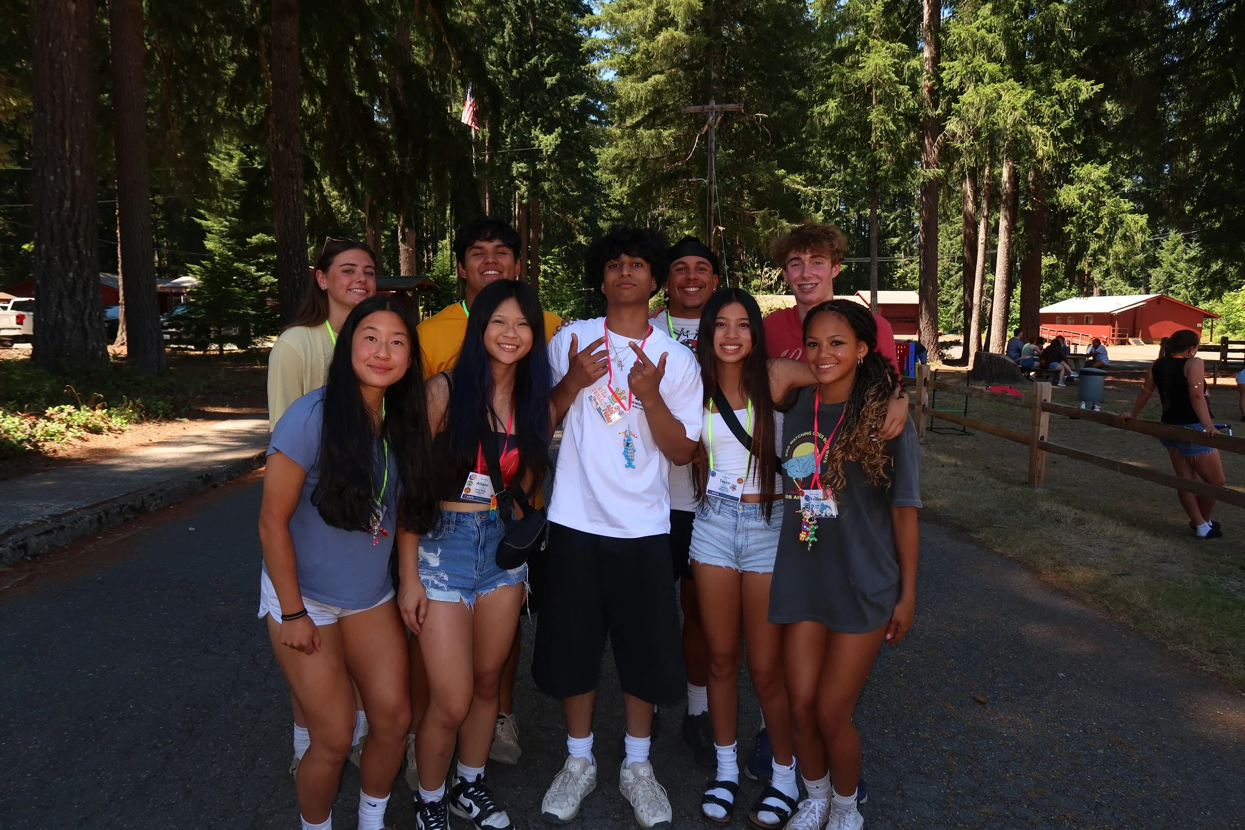 Group of nine teenagers standing outdoors in a wooded area, smiling at the camera, with some wearing lanyards and casual summer clothing.