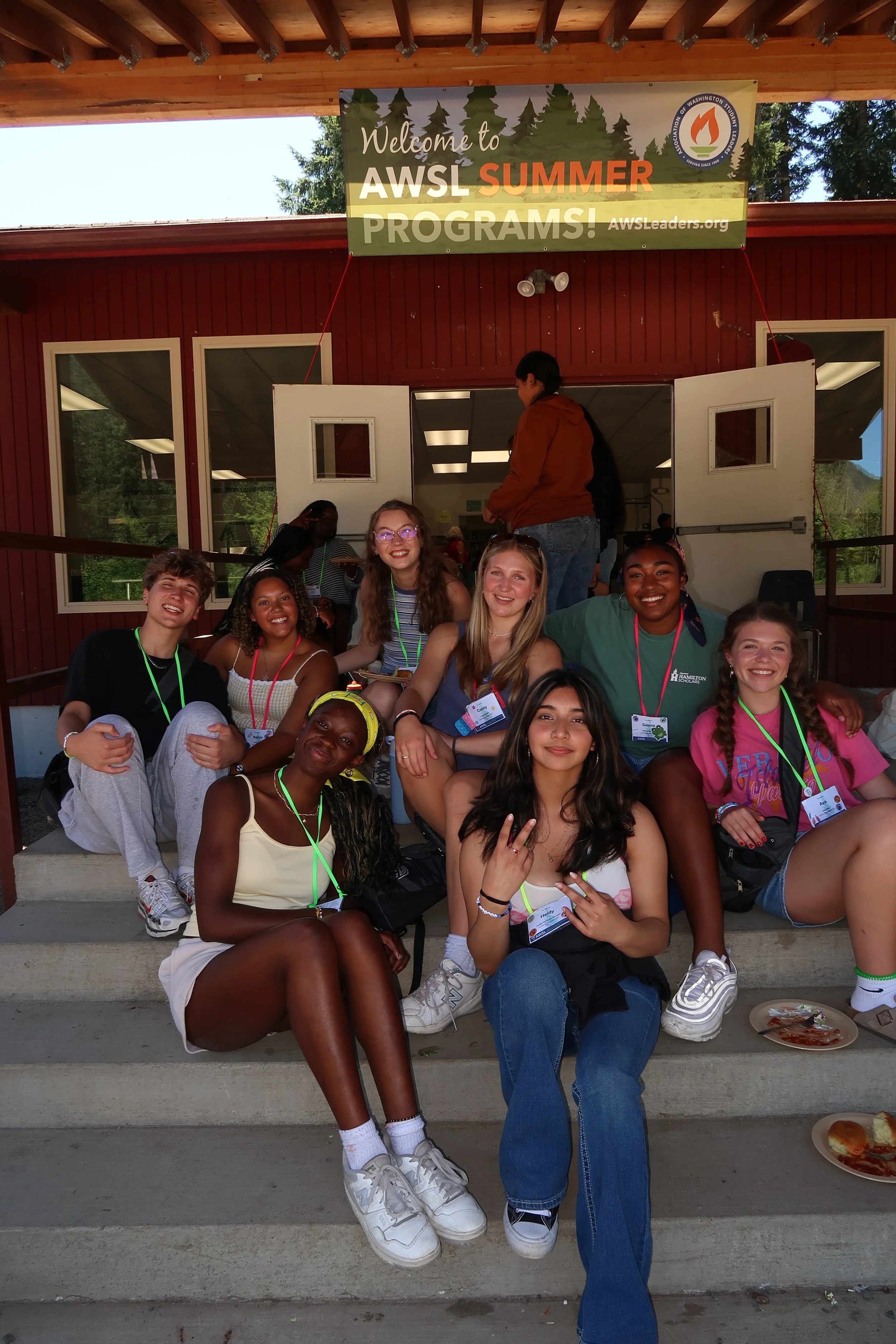 Group of young people sitting on steps in front of a red building with a sign that says 'Welcome to AWSL Summer Programs!' The group appears to be happy and enjoying each other's company, some are smiling at the camera, and some are wearing name tags