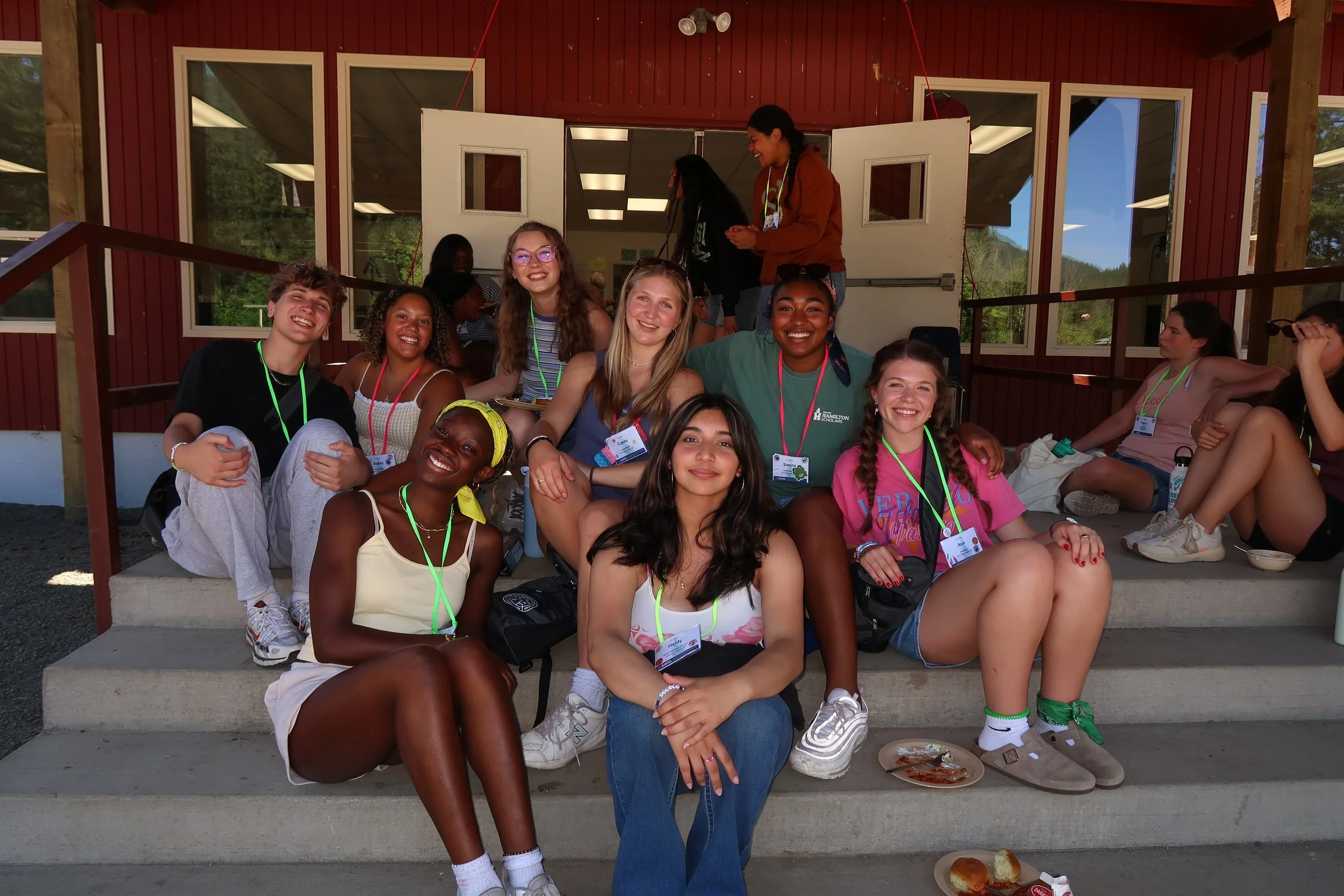 A group of twelve young people, mostly women, sitting and standing on steps outside a building with red siding and white trim, smiling and enjoying each other's company during daytime.