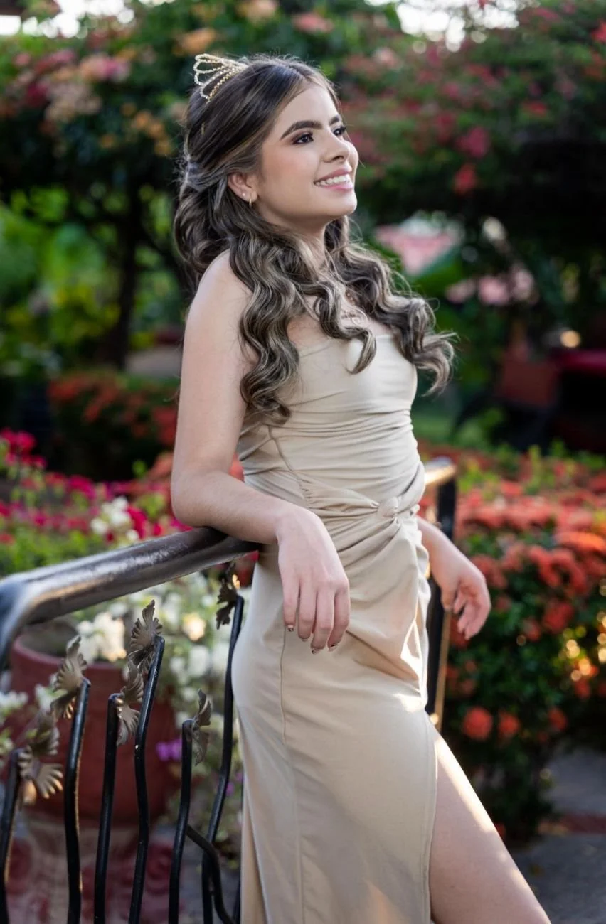 Mujer joven con vestido beige y corona de tiara, sonriendo y posando en un jardín con flores diversas.