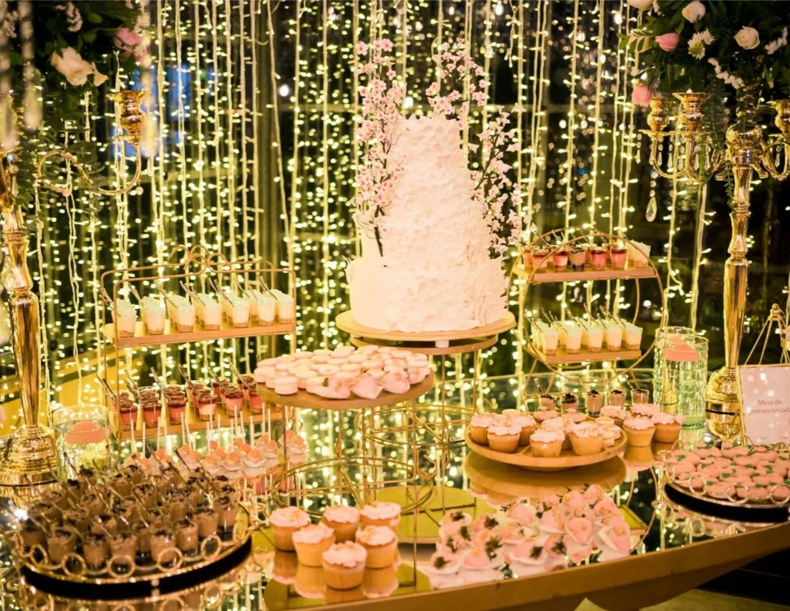 Mesa decorada con postres y pastel en un evento con fondo de luces doradas y flores.
