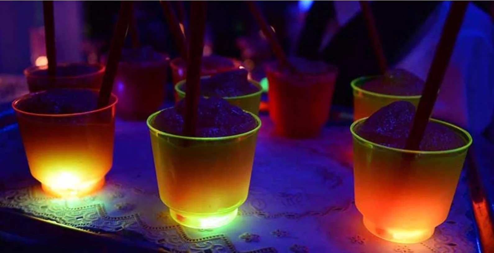 Vasos con bebidas iluminadas con luces neón en colores naranja y amarillo, con caimanes de sorbete en un ambiente oscuro.