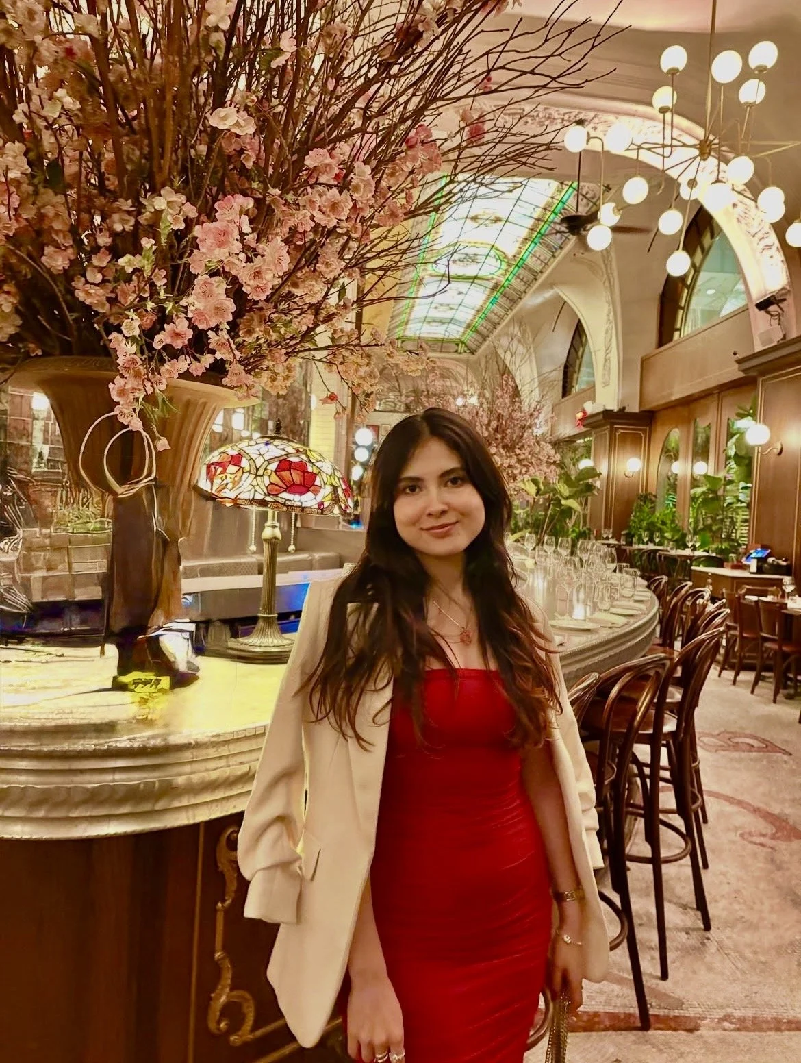 Mujer vestida con vestido rojo y blazer crema posando en un restaurante elegante con decoraciones florales y lámparas de luz cálida.