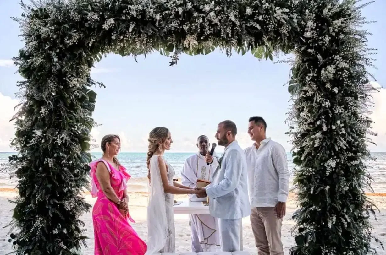 Una boda en la playa con una estructura de flores y plantas verdes. La novia y el novio se toman de las manos frente a un oficiante, mientras dos testigos observan la ceremonia.