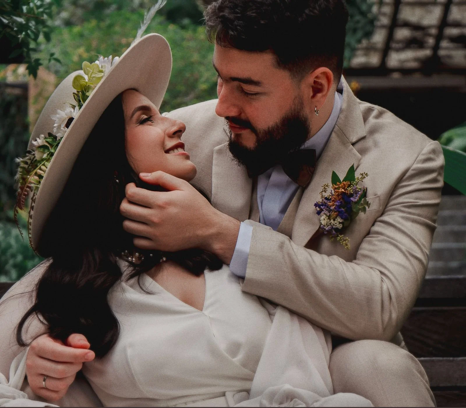 Una pareja en un momento romántico en un entorno natural. La mujer lleva un sombrero de ala ancha decorado con flores y mira sonriendo a su acompañante, quien la sostiene y la mira con cariño.